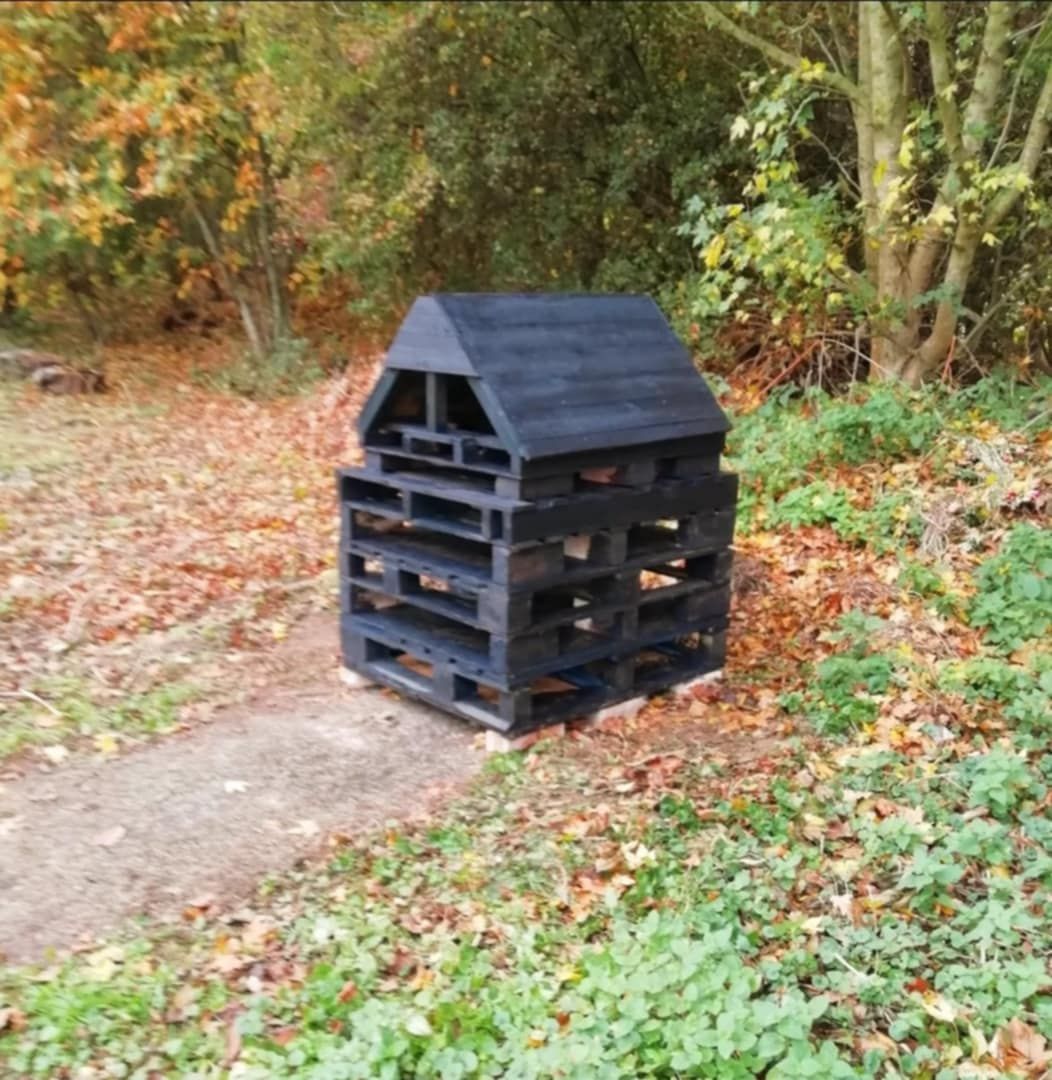 5 black painted pallets stacked on top of eachother with a black painted wooden roof on top. Bug hut is on the end of a path surrounded by leaves and trees in a church yard area